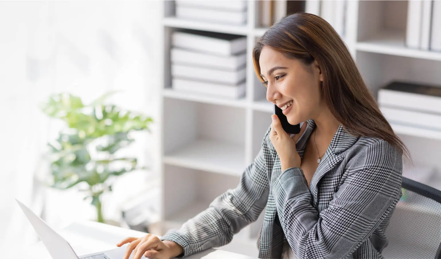 Brunette woman wearing and black and white checkered jacket talking on the phone while checking her LegalZoom virtual mail on her laptop.